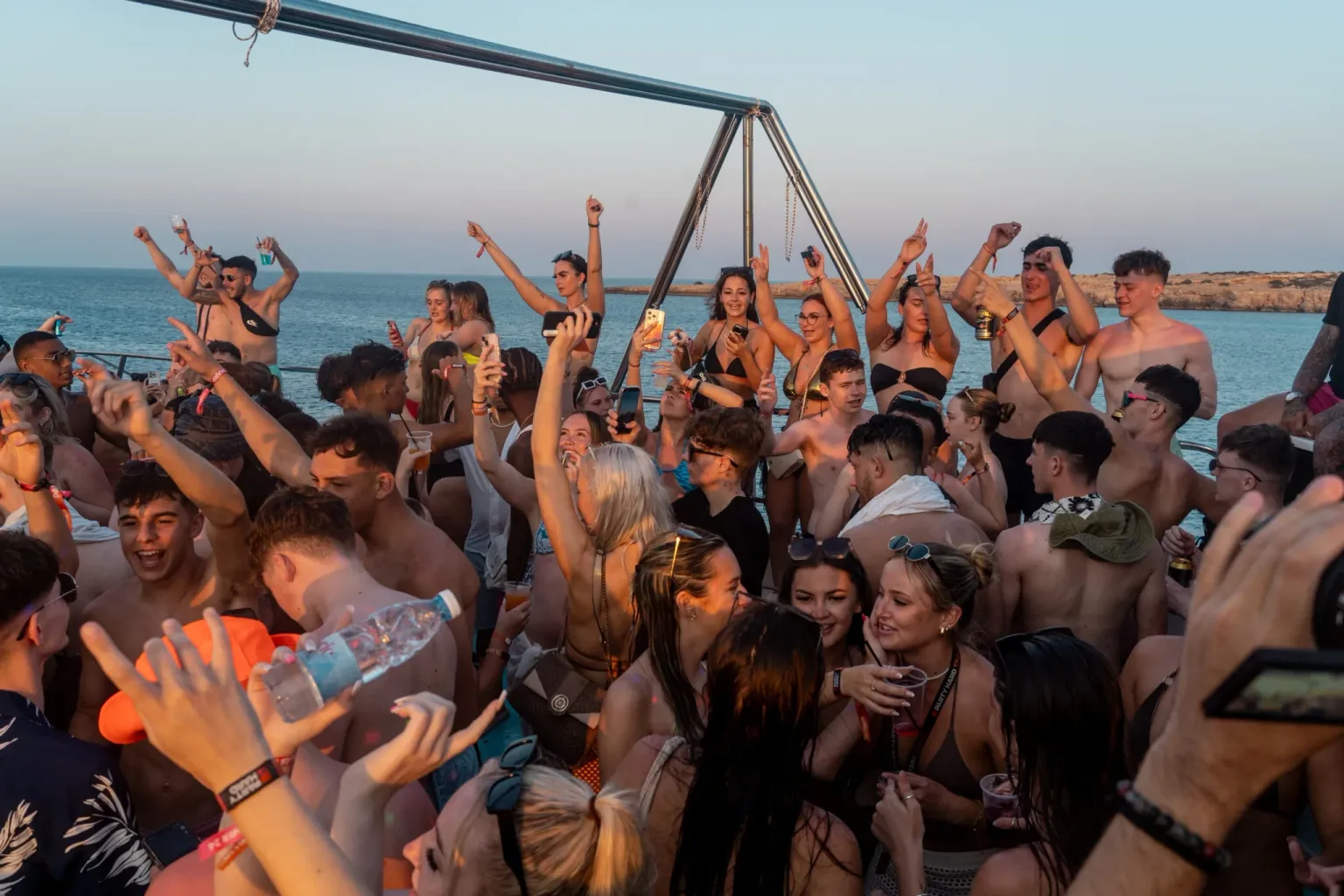 Partygoers dancing on a packed Fantasy Boat Party with the blue Ayia Napa coastline in the background