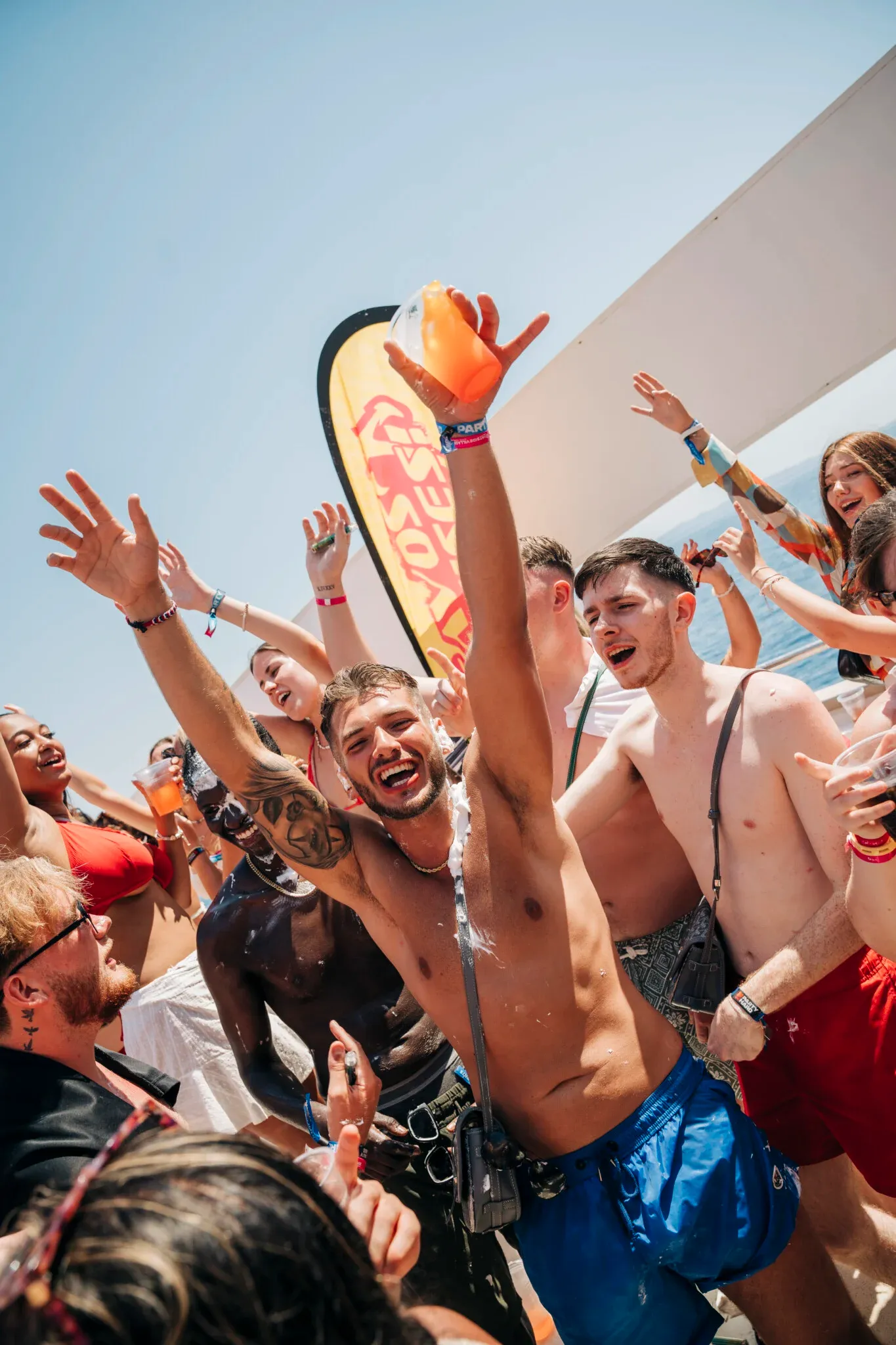 Lads and girls throw drinks in the air on the Kavos Booze Cruise, dancing on deck with hands up and party vibes going off
