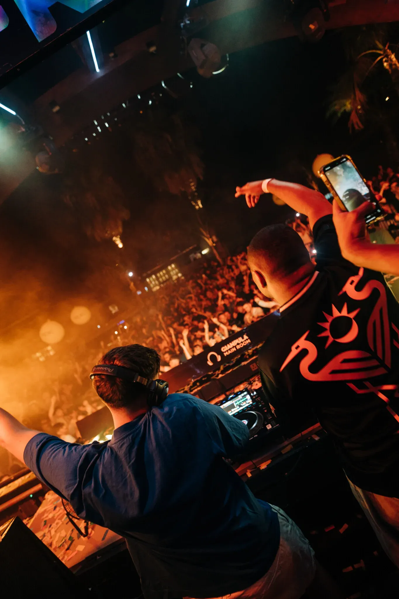 DJs performing at Gianpula Main Room Club Night in Malta, with one filming the crowd as orange smoke and stage lights fill the venue.