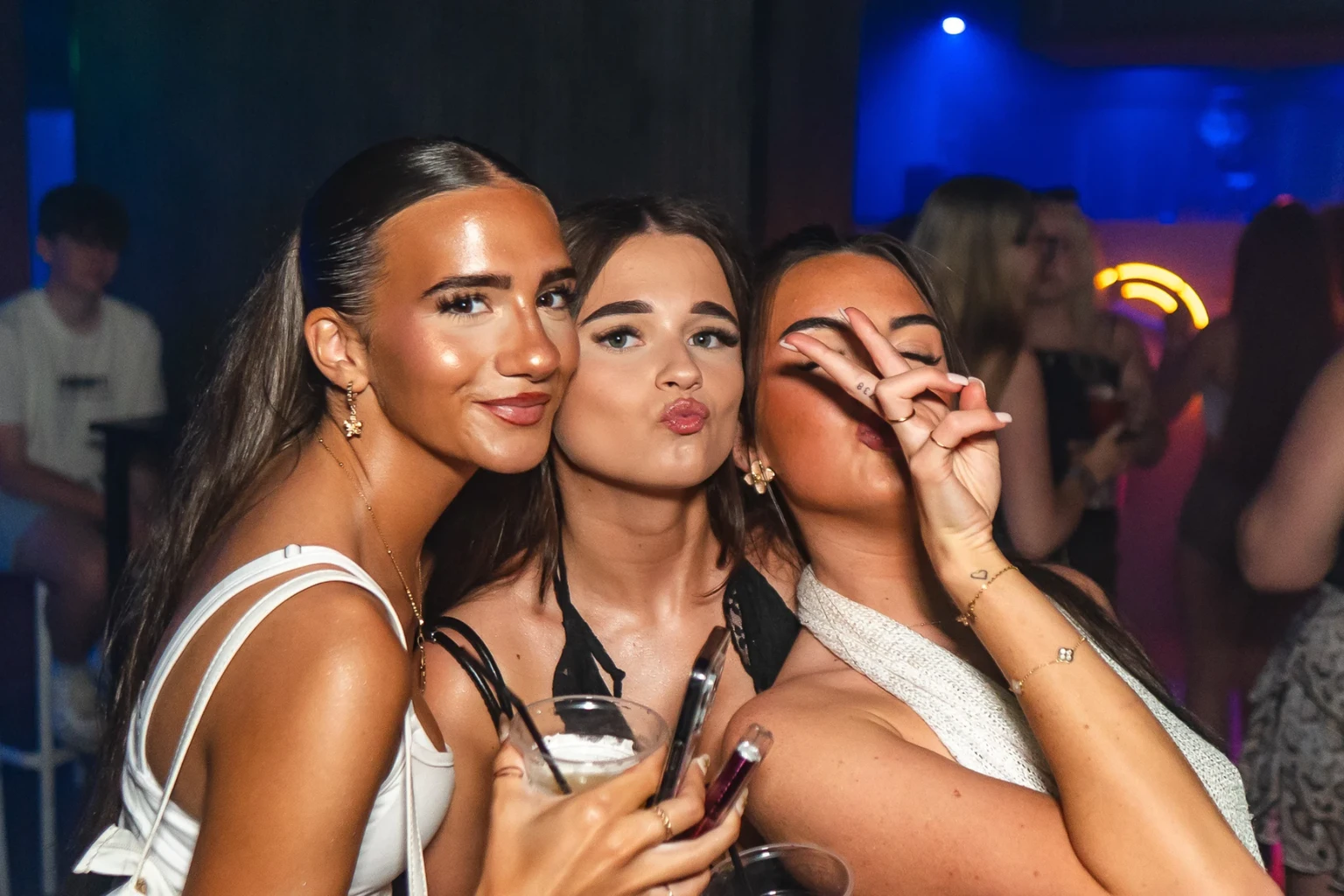 Girls posing on a packed club night, drinks in hand and buzzing with party energy under the neon lights.