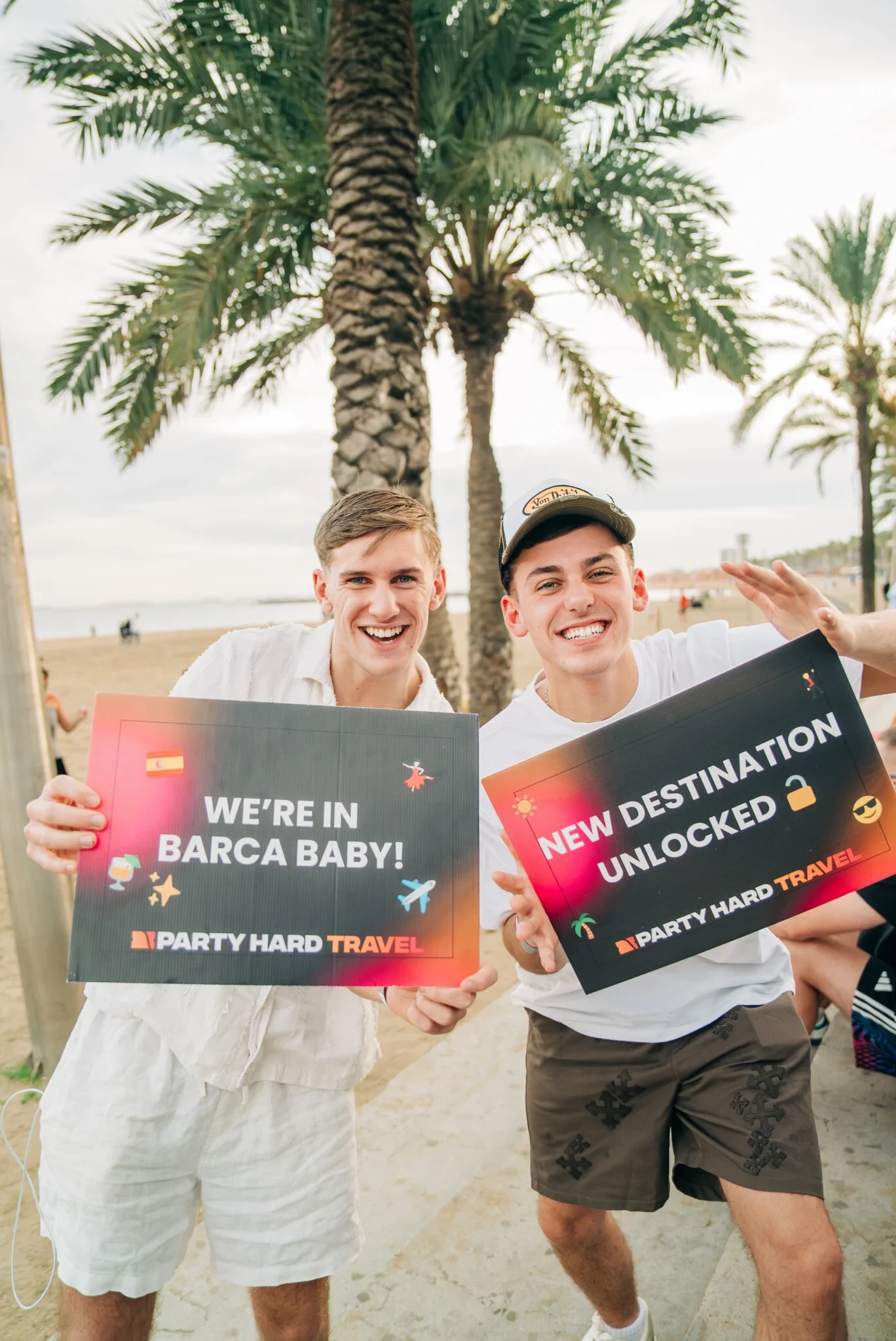 Two lads on the beach in Barcelona holding Party Hard Travel signs, buzzing for the new destination under the palm trees.