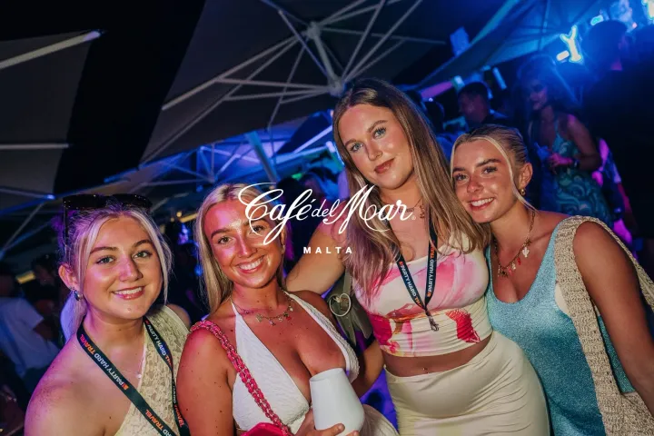 Four girls smiling at Cafe Del Mar Malta sunset party with glowing lights, DJ beats and a buzzing seaside crowd under night skies