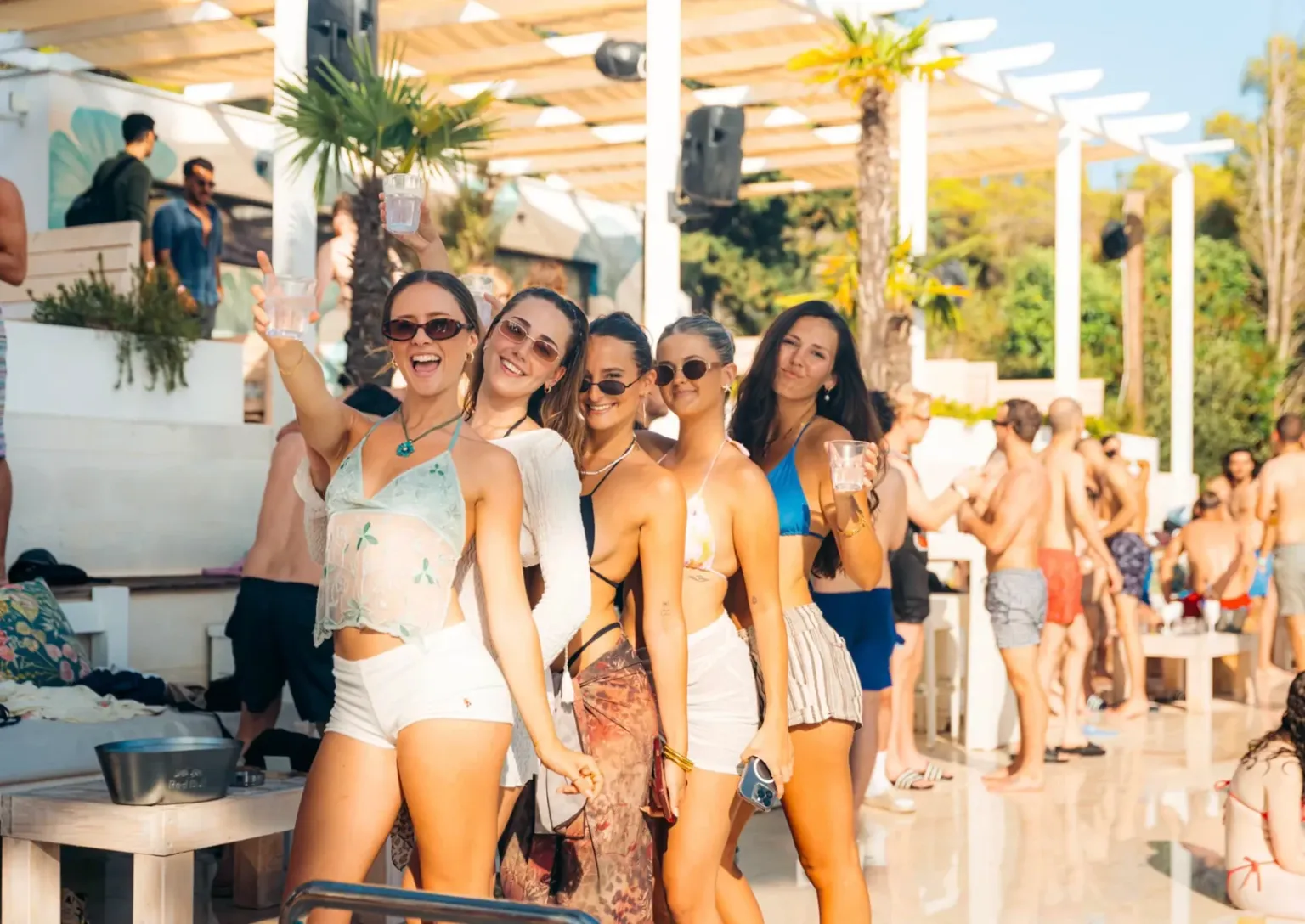 Five girls posing with drinks at the Olife Pool Party in Croatia while guests enjoy the sunshine around the poolside venue