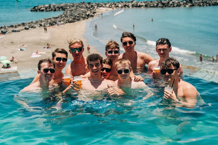 Lads packed into a Tenerife infinity pool with beers, sun out, beach below and daytime party energy in full swing