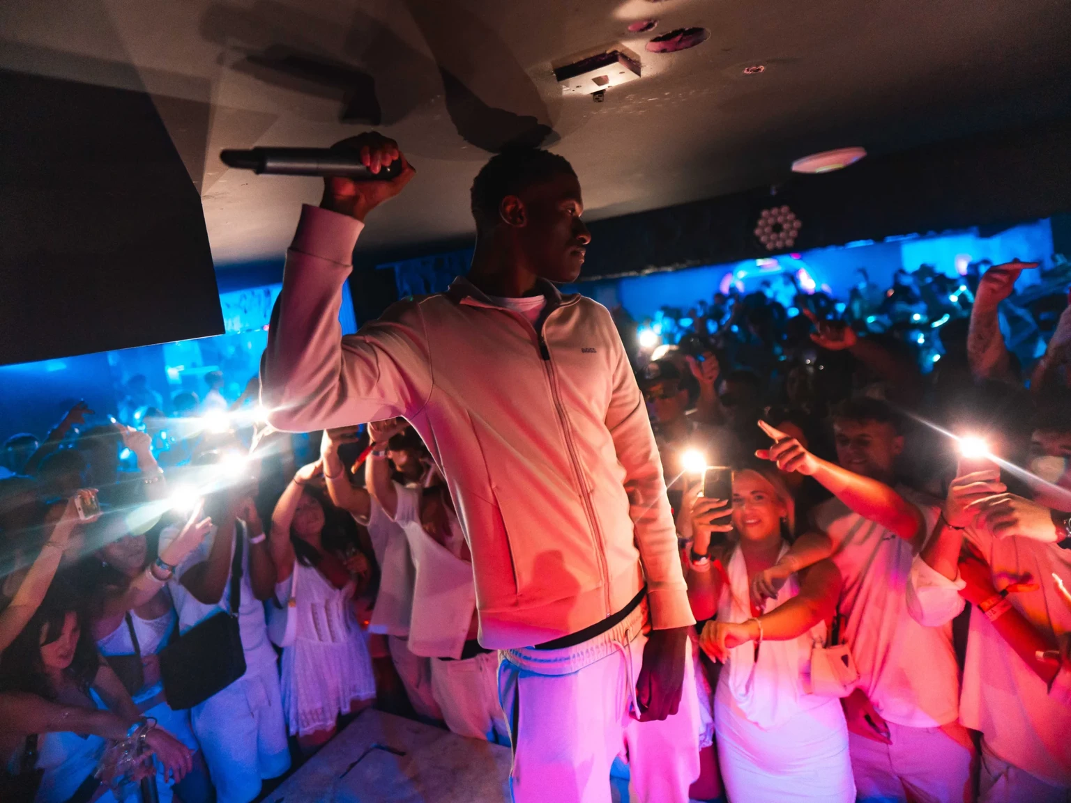MC holding a microphone on stage as the crowd in white outfits film and cheer at Napa Xtra in Ayia Napa
