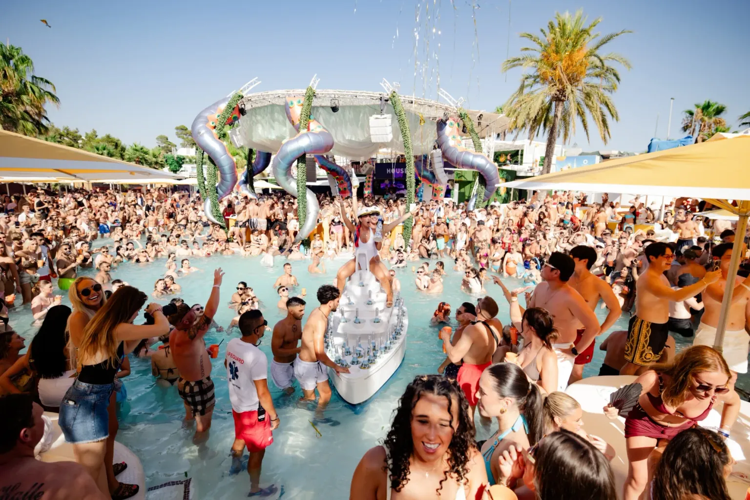 Performer standing on a drinks boat in the pool at O Beach Ibiza with a packed crowd, yellow parasols and giant tentacles above the stage