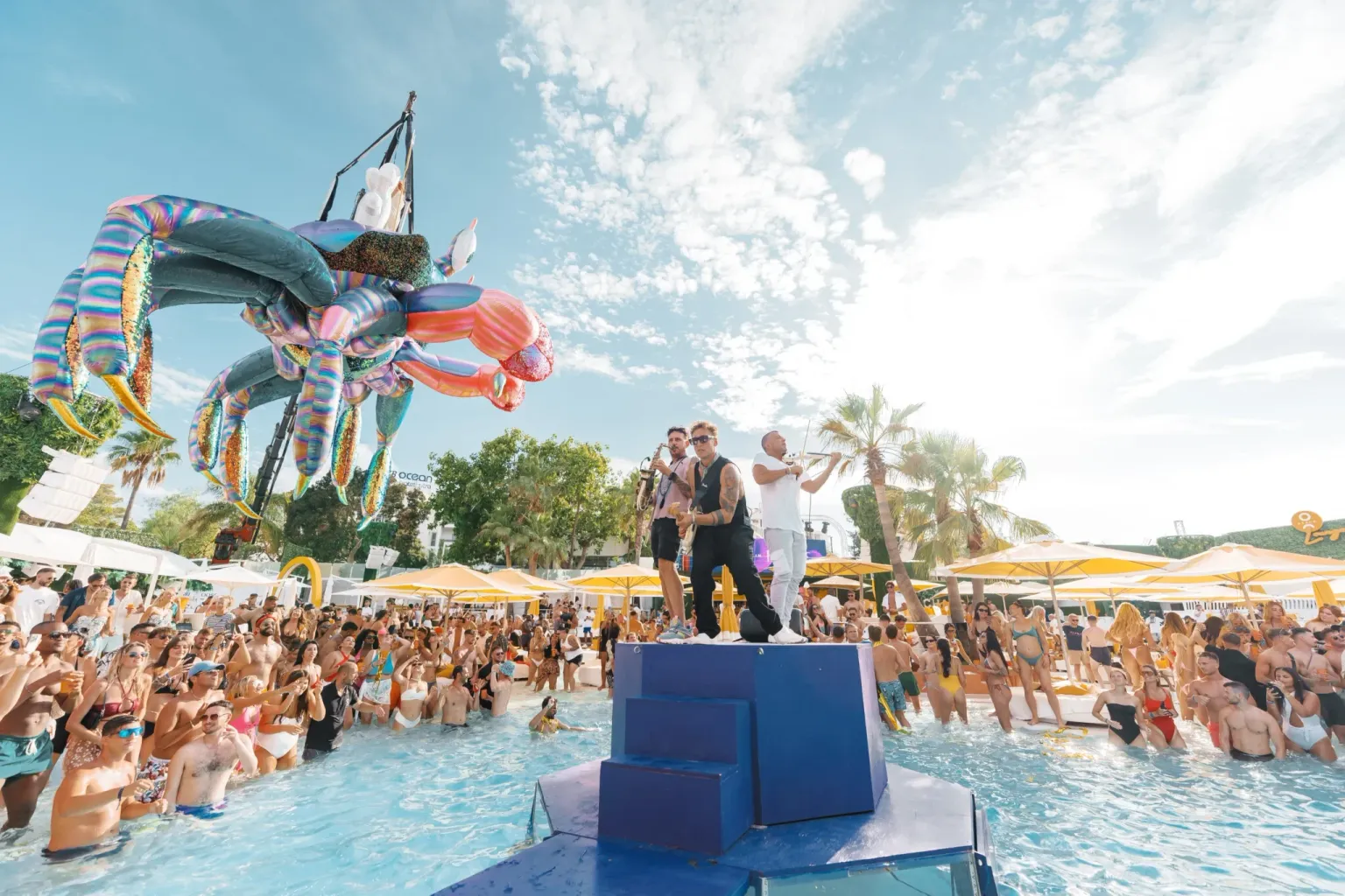 Musicians performing on a raised platform in the pool at O Beach Ibiza with a giant crab decoration above the crowd