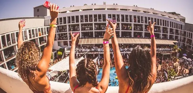 Party hard on an Ibiza clubbing holiday! Three friends raise drinks at a lively pool party with a packed club and hotel backdrop.