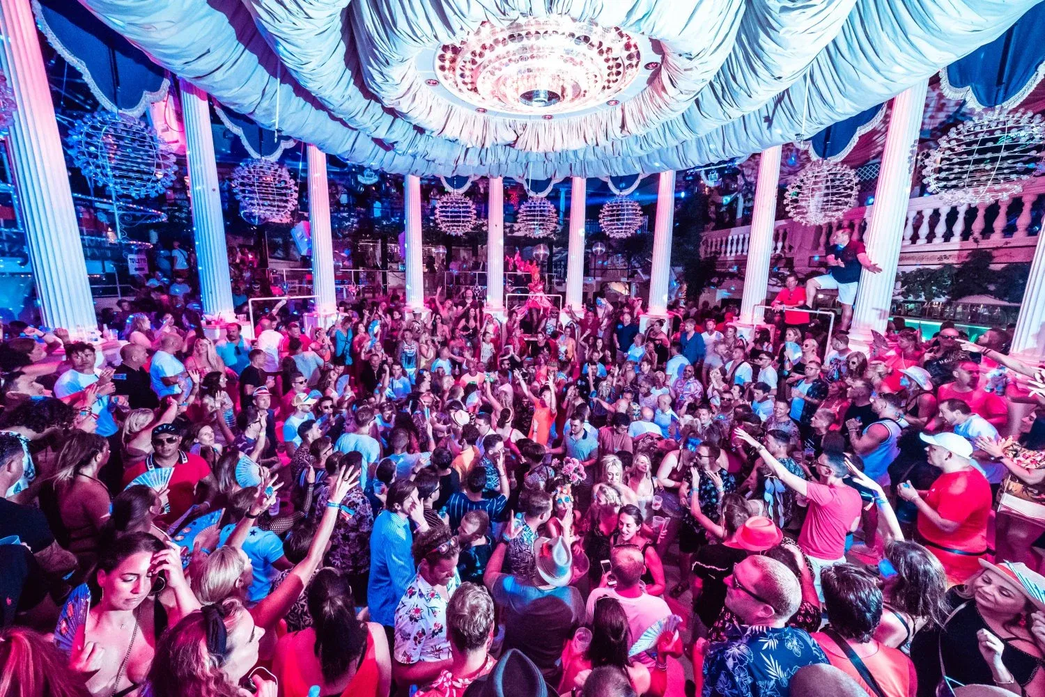 Crowd dancing inside Es Paradis Ibiza beneath chandeliers and pink and blue club lighting