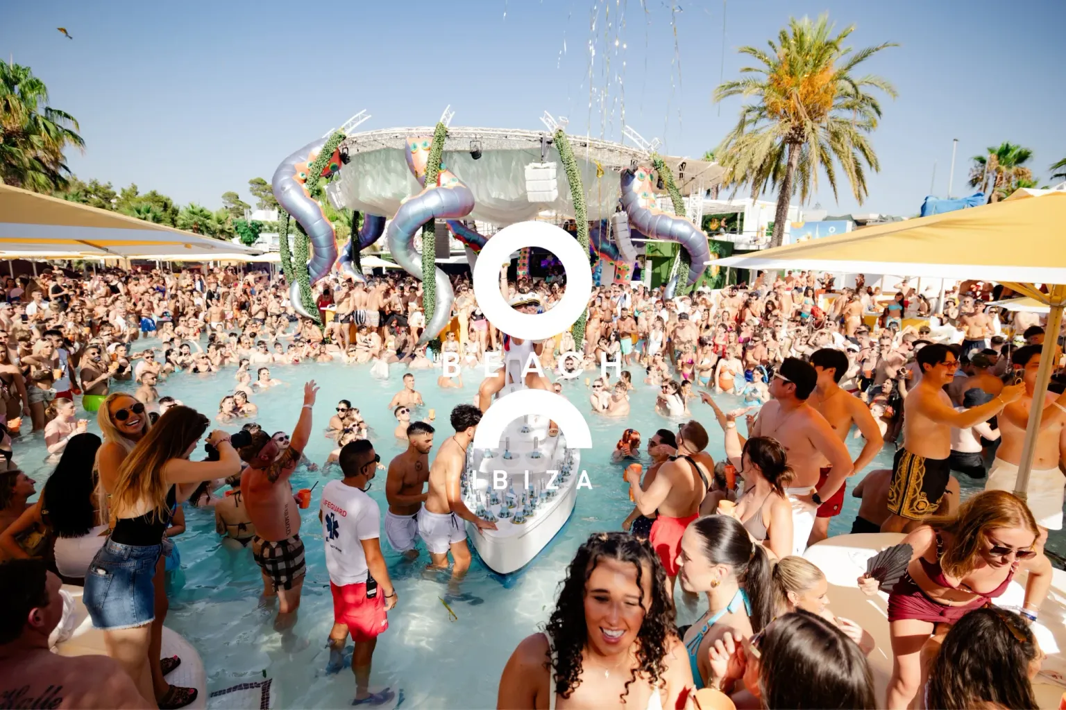 Packed crowd in and around the pool at O Beach Ibiza with palm trees, stage decor and a drinks boat in the water