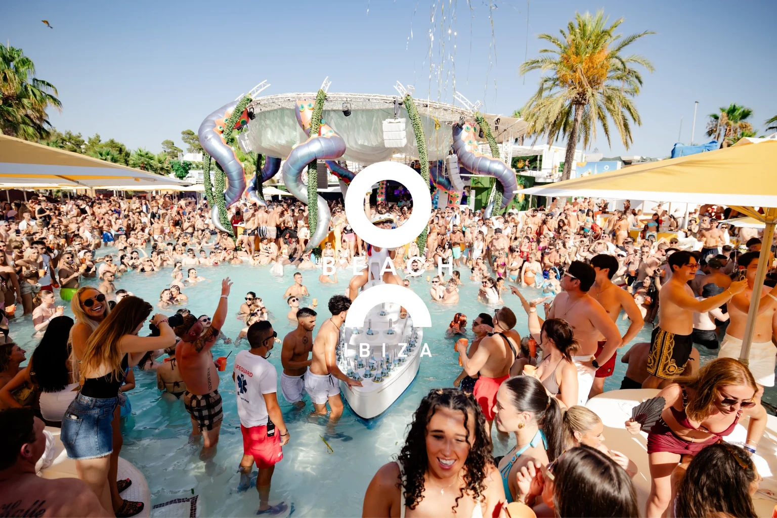 Packed crowd in and around the pool at O Beach Ibiza with palm trees, stage decor and a drinks boat in the water