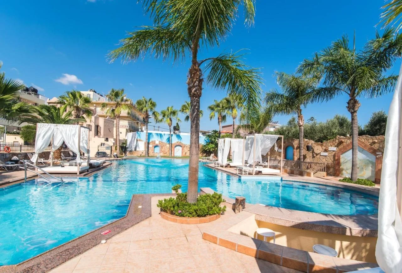 Oasis Studios Malia with palm trees, poolside beds, clear blue water and a sunny, relaxed holiday atmosphere.