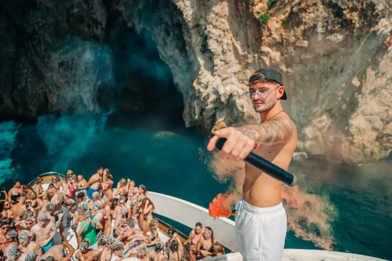 DJ holding a microphone above the crowd on the Kavos Booze Cruise near a sea cave
